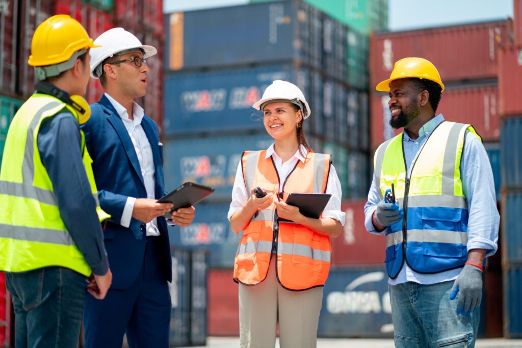 Four workers wearing safety gear and reflective vests stand together at a shipping container yard, discussing project cargo transportation with clipboards and radios in hand. Shipping containers are stacked in the background as they smile during their conversation.