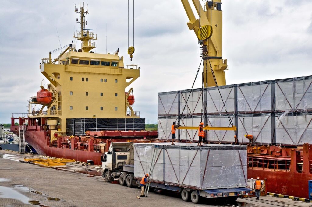 A cargo ship is docked at a port while break bulk and project cargo shipping crates are loaded onto a truck by a crane. Several workers in orange safety vests guide the operation under a cloudy sky.