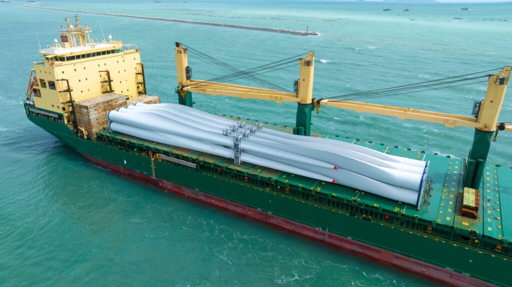 A large cargo ship specializing in break bulk and project cargo shipping transports several long white wind turbine blades on its deck across teal ocean waters, with a breakwater and smaller vessels visible in the background.