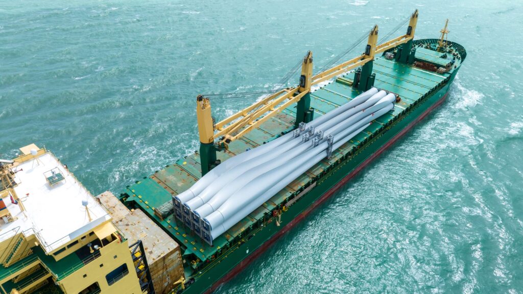 A cargo ship sails in the ocean carrying large white wind turbine blades stacked on its deck. The ship's green and yellow structures are visible amid turquoise water, highlighting the importance of marine cargo insurance for such valuable shipments.