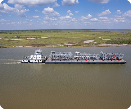 A large tugboat pushes an industrial barge carrying metal structures along a wide river, with grassy land and a partly cloudy sky in the background.