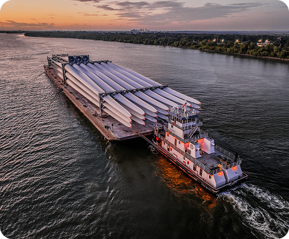 A tugboat pushes a large barge carrying multiple wind turbine blades along a wide river at sunset, with trees and distant buildings lining the riverbank.
