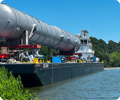A large industrial vessel carries a long cylindrical tank along a river, with trees and blue sky in the background. The tank is secured on the barge, and various equipment is visible on deck.