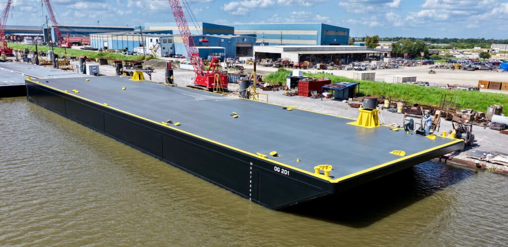 An empty black and grey barge labeled “OG 201” docked at an industrial waterfront.
